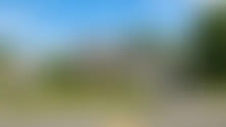 Two-story suburban house with beige siding, dark roof, green lawn, and driveway. Trees and clear blue sky in the background. 