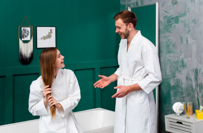 A man and woman in white robes talk happily in a modern bathroom with green walls. She brushes her hair near a bathtub.