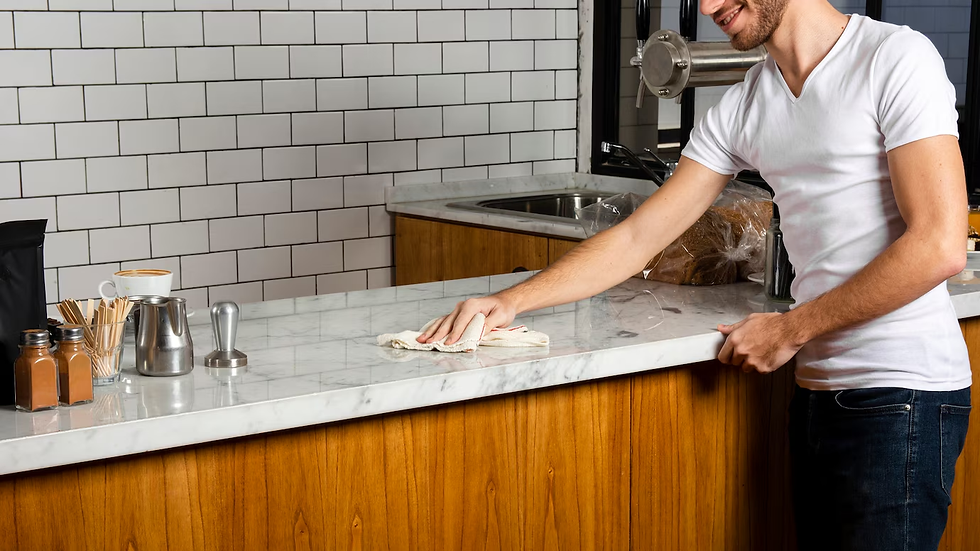 Man cleaning a marble countertop in a cafe with a tiled wall behind. Coffee items visible. Bright and tidy setting.