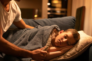 Child sleeping on a couch, covered with a blue blanket. An adult tucks them in. Warm lighting creates a cozy, peaceful atmosphere.