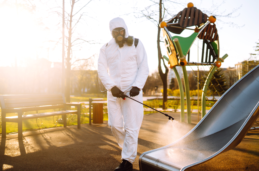 A person in a white hazmat suit disinfects a playground slide, with morning sunlight casting long shadows. Trees and benches in the background.