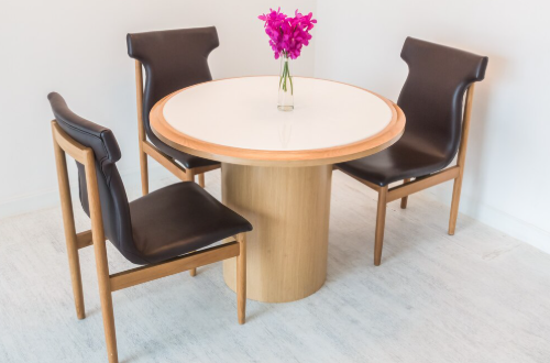 Round wooden table with a white top, three dark brown chairs, and a vase of bright purple flowers on a light wood floor.