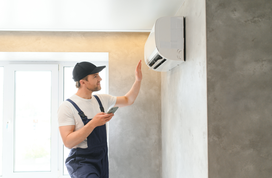 Man in blue overalls and cap holds a phone, checking an air conditioner on a gray wall. Bright room with a large window. Focused mood.