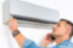 Man in denim shirt examines air conditioner while on phone, adjusting its panel. White wall background, focused and attentive.