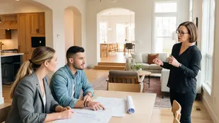 A woman in black presents to a seated couple reviewing blueprints in a bright, modern living room with wooden decor, conveying a professional mood.
