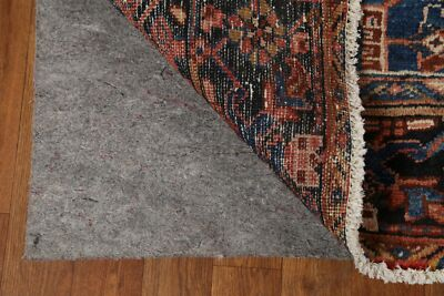 Corner of a patterned rug lifted to reveal a grey rug pad beneath, on a wooden floor. The rug has intricate blue, red, and beige designs.