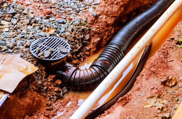 Black drainage pipe in muddy trench, with gravel and brown soil around. Water visible at the base, creating a damp, earthy atmosphere.