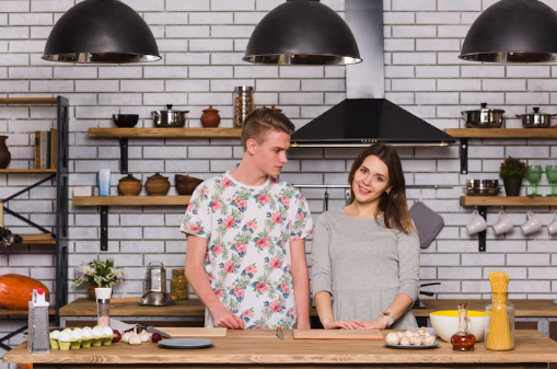 Two people stand in a kitchen with brick walls. The man wears a floral shirt, glancing at the woman in gray. Cooking ingredients are on the counter.