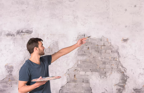 Man painting on a textured wall with a brush and palette, wearing a navy shirt. Gray background with exposed brick details. Concentrated mood.