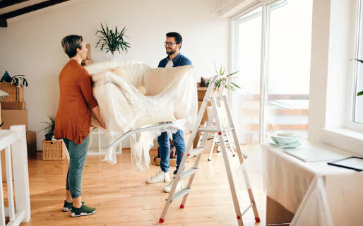 Two people unpacking a large item wrapped in plastic in a bright room with wooden floors, plants, and boxes. There's a ladder beside them.