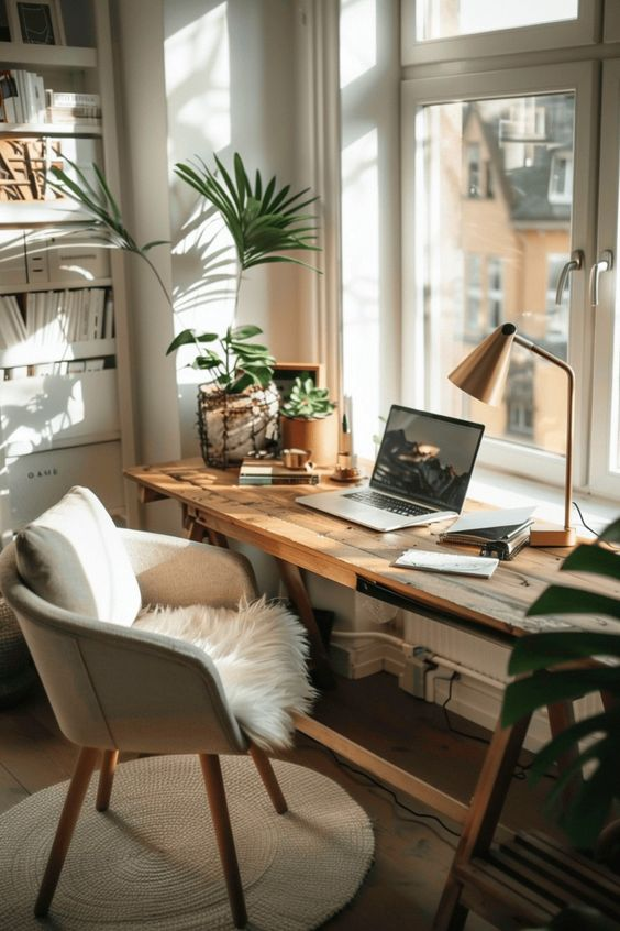 A cozy workspace with a wooden desk, laptop, lamp, books, and plants by a sunlit window. Beige chair with a fur throw adds warmth.