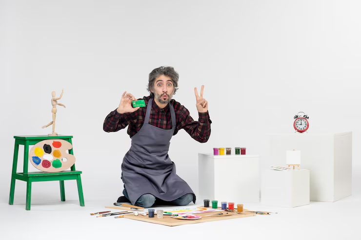Man in apron kneels on a floor with paints and brushes, holding green paint and making a peace sign. White background, colorful art scene.