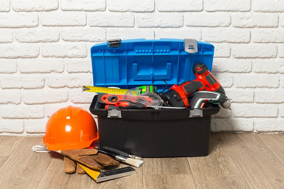Toolbox with drills, tape measure, gloves, and an orange hard hat on wooden floor. Background: white brick wall.