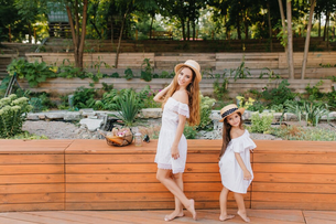 Mother and daughter in white dresses and straw hats pose happily in a garden with wooden steps, lush greenery, and a basket of fruit nearby.