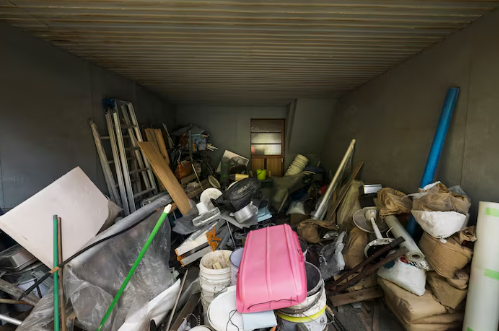 Cluttered storage room with stacked tools, furniture, pink object, and bags. Gray walls and scattered items create a chaotic atmosphere.