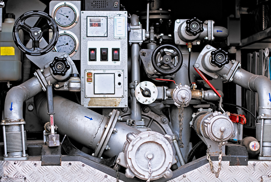Complex machinery with pipes, gauges, and valves in a metallic setting. Dials show numbers and arrows; mood is industrial and technical.