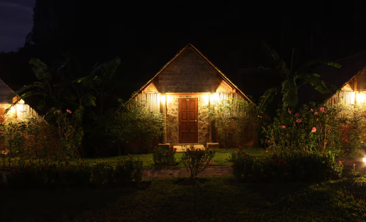 Triangular cabin with wooden door lit warmly at night. Surrounded by plants and flowers. Peaceful atmosphere with dark sky.