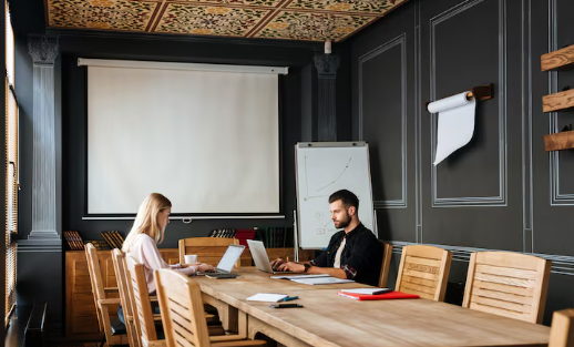 Two people work on laptops in a modern conference room with wooden chairs. There are a projector screen, a whiteboard, and dark walls.