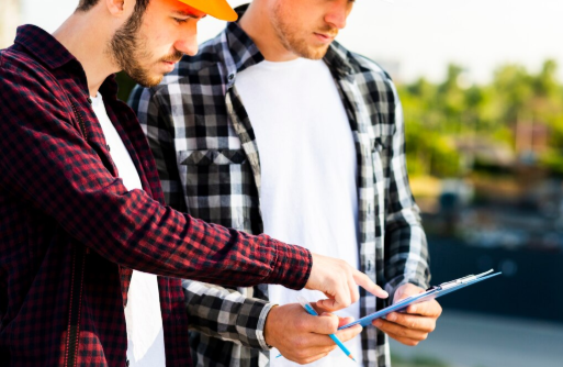 Two men in plaid shirts and hard hats discuss blueprints outdoors. One points at the clipboard. Background shows blurred greenery.