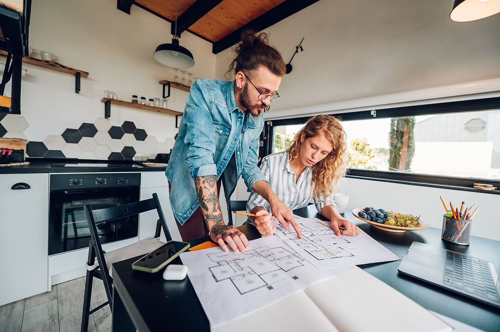 Two people in a modern kitchen study house plans on a table. Hexagonal tile backsplash, fruit bowl, and laptop visible. Focused mood.