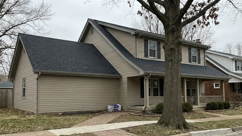 Two-story beige house with a gray roof, front porch, and large bare tree. Lawn has scattered leaves and stacked bags near the entrance. Cloudy sky.