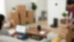 Two women in a living room; one on a laptop with a calendar, another packing boxes. Pile of labeled boxes, plants, tablet on table, cozy setting.