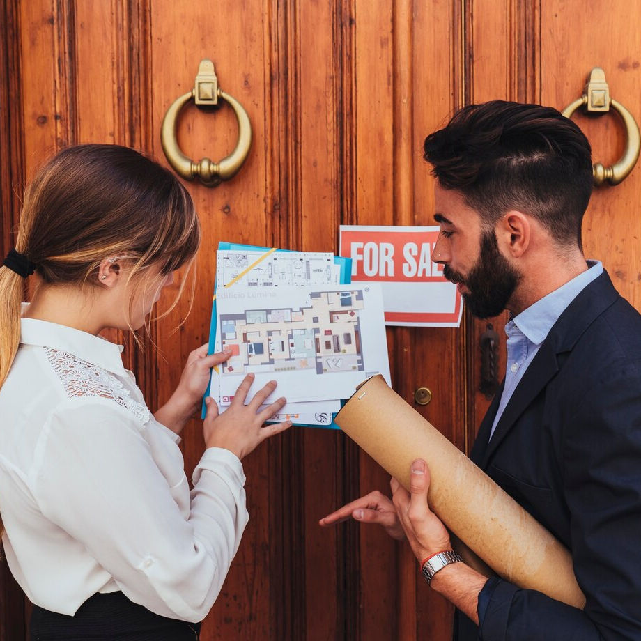 Two people discuss floor plans outside a wooden door with a "For Sale" sign. One holds rolled-up papers, both focused on plans.