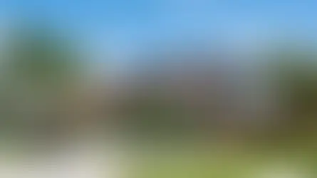 Modern two-story house with a beige exterior, brown tiled roof, and solar panels. Surrounded by palm trees and lush greenery, under a clear blue sky.