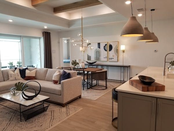 Modern living room and kitchen with neutral tones, beige sectional couch, stylish lighting, abstract art, and wood accents on counters. Cozy mood.