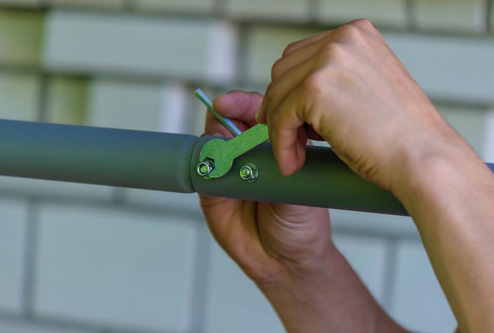 Hands tighten bolts on a grey metal pipe using a green wrench. Background shows blurred light-colored bricks.