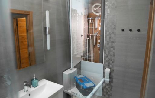 Modern bathroom with gray tiles, a sink with soap, glass shower door, and blue vacuum cleaner. Pipes and fittings visible behind glass.