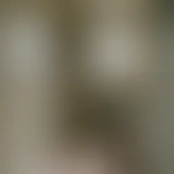 Small bathroom with a shower, sink, and toilet. Beige tiles, floral-patterned floor, and a window. Soap and bottles on shelves.