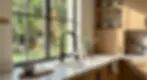Sunny kitchen with a black faucet, wooden cabinets, a plant, cups, and utensils. Greenery visible outside the large window. Cozy feel.