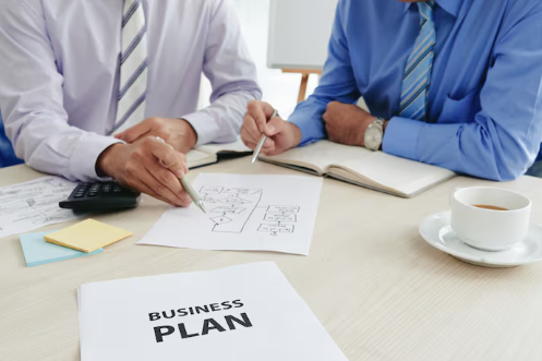 Two people in shirts discussing documents on a table with a "BUSINESS PLAN" paper, coffee cup, and notebook. Office setting, focused mood.