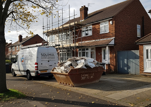 White van and dumpster with debris outside a brick house under renovation. Scaffold on facade. Tree on left, pavement with fallen leaves.