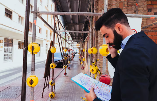Man in suit on phone, holding blueprints at construction site. Yellow safety caps line scaffolding. Urban street in background.