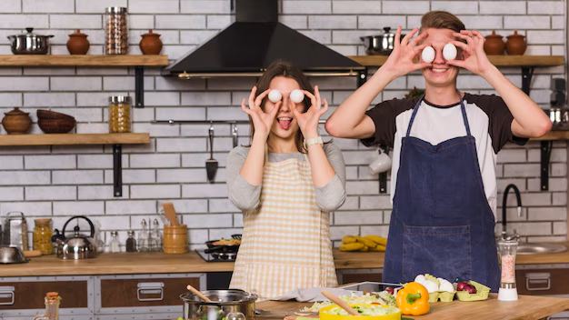 Two people in aprons playfully hold eggs to eyes in a kitchen. Background features shelves, utensils, and white brick walls.