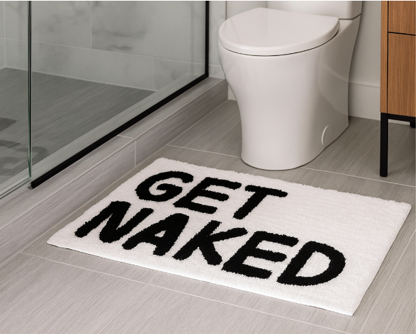 White bathroom mat with "GET NAKED" in bold black letters on gray tile floor, next to a toilet and glass shower. Modern, humorous vibe.