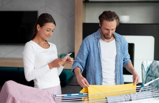 A man folds a yellow shirt on a drying rack. A woman beside him smiles at her phone. They're in a modern living room with soft lighting.