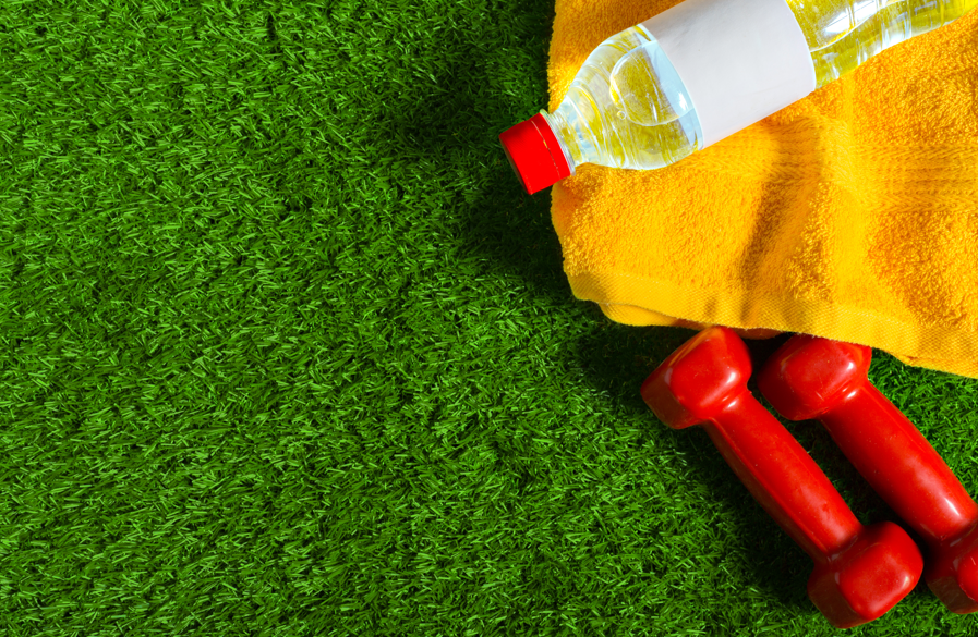 Red dumbbells, a yellow towel, and a water bottle lie on green grass. The setting is vibrant and sporty. No visible text.