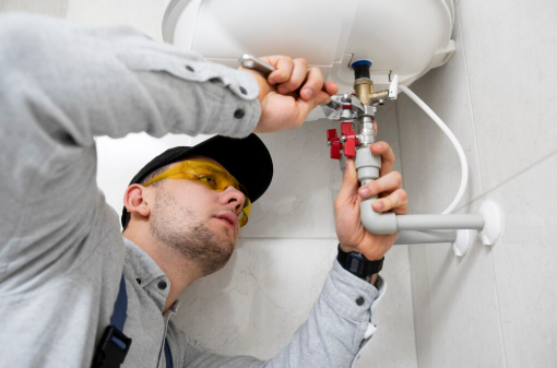 A plumber in grey clothes and yellow safety glasses works on pipes under a sink in a tiled bathroom. Focused expression.