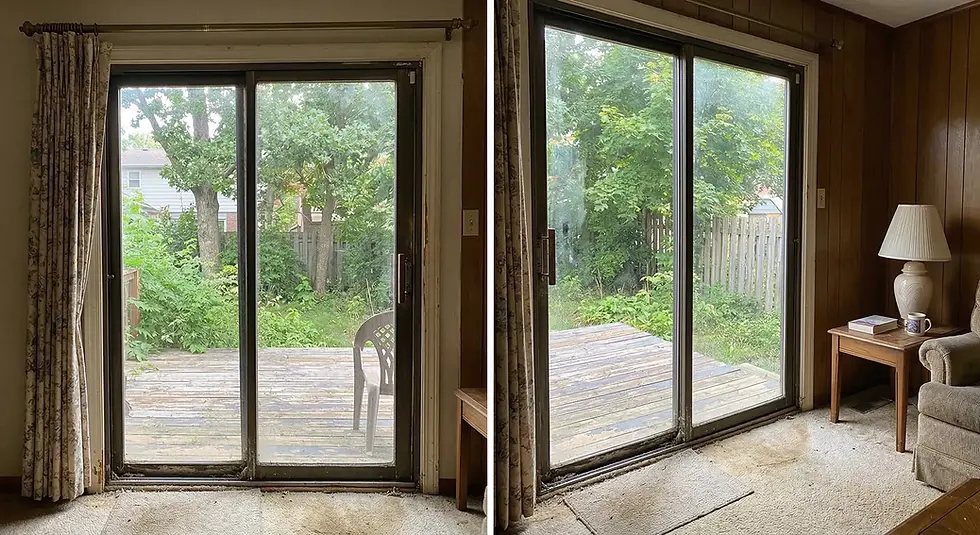 Sliding glass doors open to a wooden deck, overgrown garden, and trees. Indoors: floral curtains, a lamp on a table, and beige carpet.