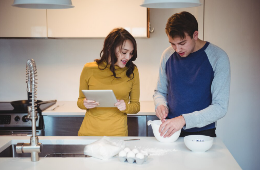 A woman with a tablet and a man mixing ingredients are in a modern kitchen. They are smiling, surrounded by eggs and flour.