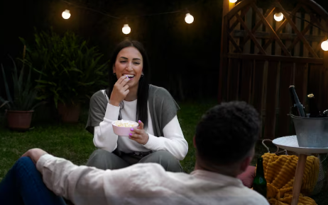 Woman smiling, eating popcorn outdoors at night. String lights illuminate the yard with plants and a table with drinks, creating a cozy mood.