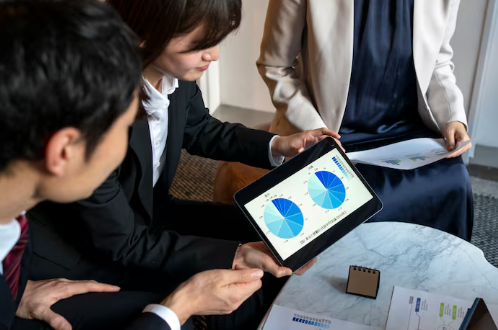 Three people in business attire discuss charts on a tablet in an office setting. Papers on the table show pie charts. Professional mood.