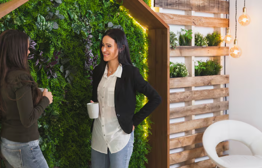 Two women chat holding mugs by a vertical garden, indoors. Warm lighting, wood accents, and white chair create a cozy atmosphere.