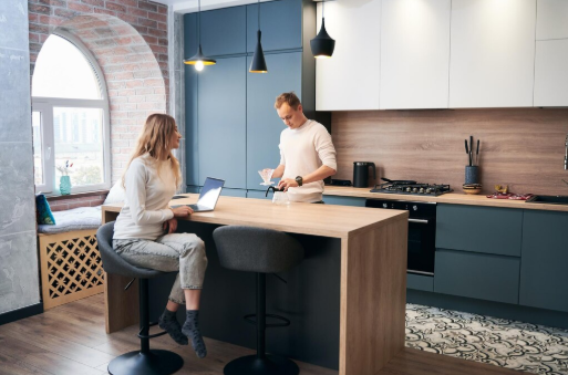 Woman with laptop sits at kitchen island talking to standing man. Modern kitchen with blue cabinets, wood accents, and large window.