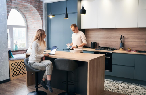 A man and woman in a modern kitchen, the woman seated with a laptop and the man pouring a drink. They appear relaxed and engaged.
