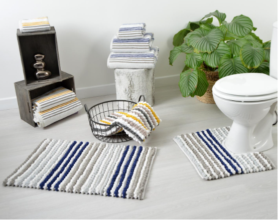 Striped bathroom rugs in blue, gray, and beige near a white toilet. Towels stacked on shelves, plant in the background, serene setting.