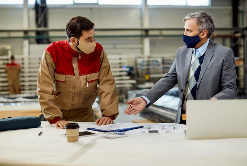 Two men in masks discuss blueprints on a table in an industrial setting. One wears a red work jacket, the other a gray suit. Mood is focused.
